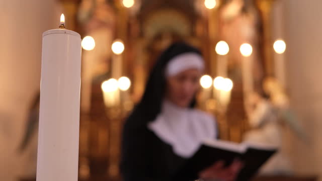 Burning white candle in front focus, nun reading Holy Bible in Roman Catholic church in the background.