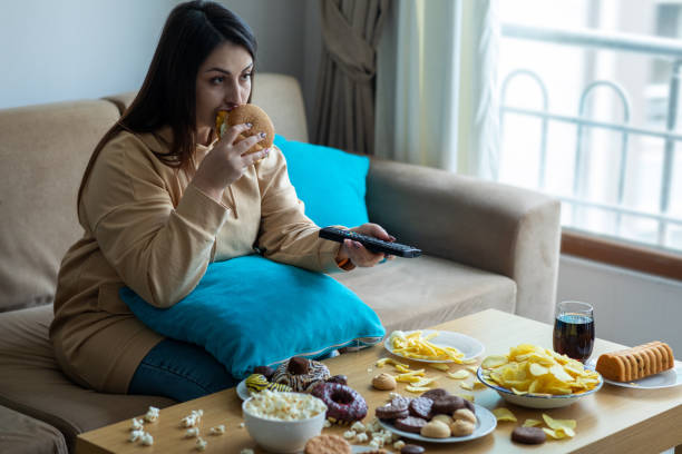 mujer con sobrepeso sentada en el sofá - comer demasiado fotografías e imágenes de stock