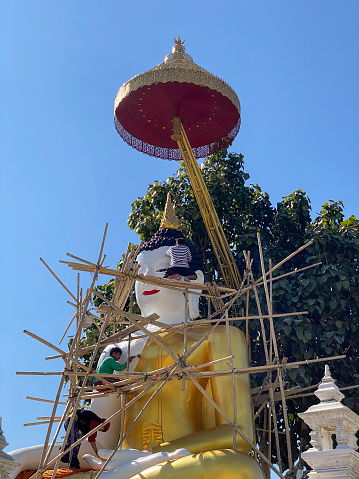 This newly finished constructed concrete Buddha image is carefully painted by construction workers on the grounds of the temple. Once the painting is complete and the scaffolding removed an 'Opening of the eyes' ceremony will be enacted to imbue spirit into the sacred Buddha image. These kinds of Buddhist ceremonies are familiar across the Kingdom of Thailand regularly when a Buddhist statue is constructed and completed.