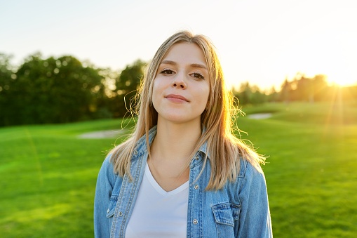 Portrait of smiling beautiful happy young woman 20 years old, sunset green lawn in park background