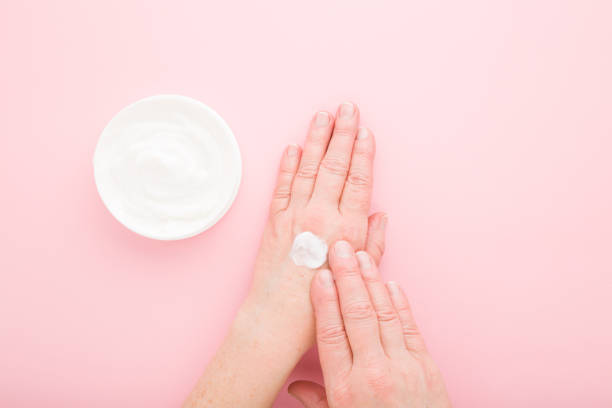 istock Mature woman hands using white anti aging moisturizing cream on light pink table background. Pastel color. Care about soft body skin in old ages. Daily beauty product. Closeup. Top down view. 1300822941