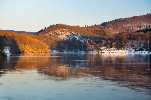Evening view over the frozen Obersee of lake Rursee with reflection of the illuminated landscape in winter in the Eifel, Germany.