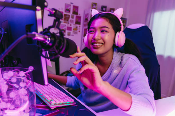 Excited and smiling gamer girl in cute headset with mic playing an online video game. Young Asian woman talking to players and audience on personal computer at home Excited and smiling gamer girl in cute headset with a mic playing an online video game and live streaming. Young Asian woman talking to players and audience on computer in neon led light room at home streamer stock pictures, royalty-free photos & images