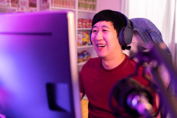 aufgeregt und lächelnde männliche spieler in headset mit mikrofon spielen ein online-videospiel. junger asiatischer mann im gespräch mit spielern und publikum auf pc zu hause - nerd computer learning fun stock-fotos und bilder