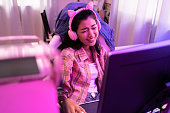 Excited and smiling gamer girl in cute headset with mic playing an online video game. Young Asian woman talking to players and audience on personal computer at home