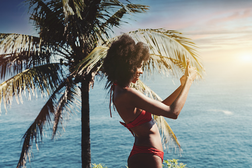 A cheerful young African-American female with curly hair and in swimwear is having an online vlog broadcast via her smartphone while standing on the ocean coastline of a tropical resort near the palm
