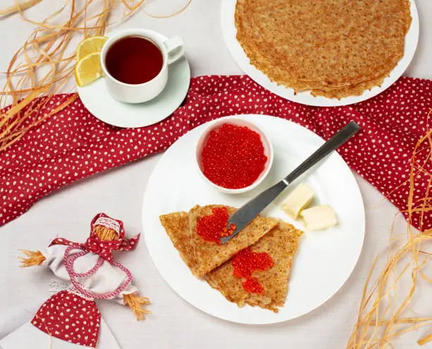 Pancakes with red caviar on the white plate and traditional symbol scarecrow winter. Staple of yeast pancakes, traditional for Russian pancake week. Shrovetide or Maslenitsa, Butter Week festival meal. Close up top view