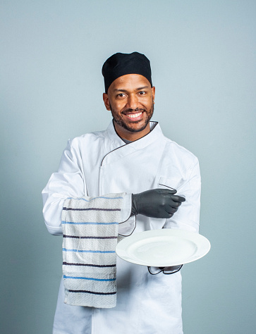 Happy male cook holding napkin and empty plate while serving. Portrait of confident chef in white uniform standing against gray background.