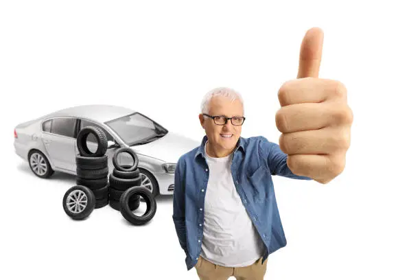 Mature man standing in front of a silver car and piles of tires, showing thumbs up isolated on white background