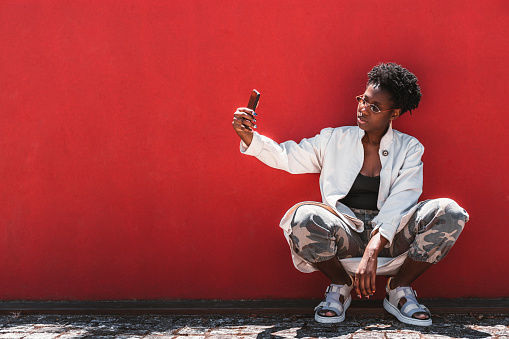 Portrait of a stylish young black female with curly Afro hair, in spectacles, white trench, camouflage trousers, taking a selfie via cellphone while squatting near a solid red wall; a copy space area