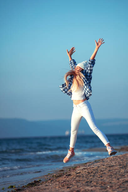young beautiful woman, wearing blue shirt and white jeans, having fun jumping on the beach, near the sea, hands in the air,