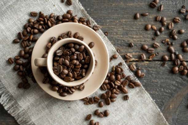 신선한 아라비카 또는 로부스타 커피 콩이 린넨 섬유와 목재 테이블에 흩어져 있는 커피 원두로 채워진 커피 컵. - latté cookie cappuccino coffee crop 뉴스 사진 이미지