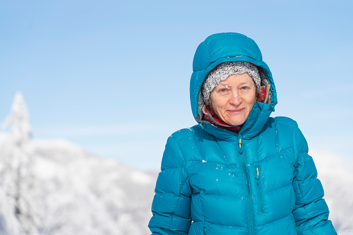 Mature cheerful women in blue winter parka, waist up, against clear blue sky looking at camera, sunlight.