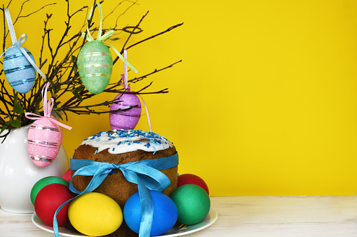 Easter bouquet of dry twigs in a jug and festive cake with colorful painted eggs on yellow and white background