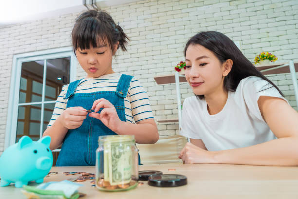 Mother is teaching daugther on financial saving and planing using saving jar and piggy bank with real moner, for Money and wealth education concept. stock photo