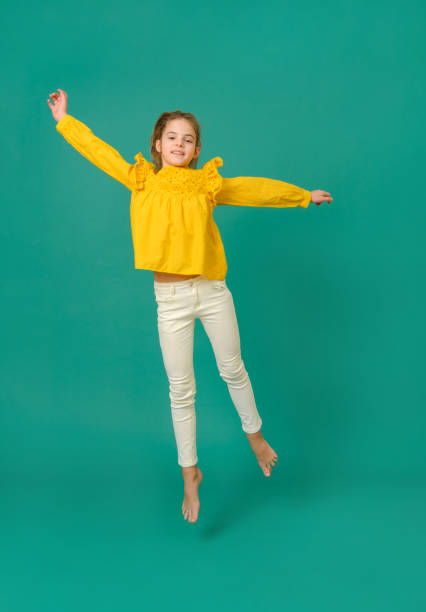 ragazza adolescente bianca di 10 anni in una camicetta gialla e pantaloni bianchi salta su uno sfondo verde - 10 11 years cheerful happiness fun foto e immagini stock