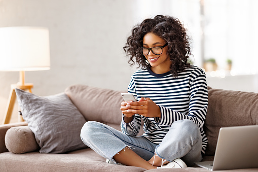 Delighted ethnic woman in casual clothes and glasses smiling and browsing social media on smartphone while sitting cross legged on sofa near laptop