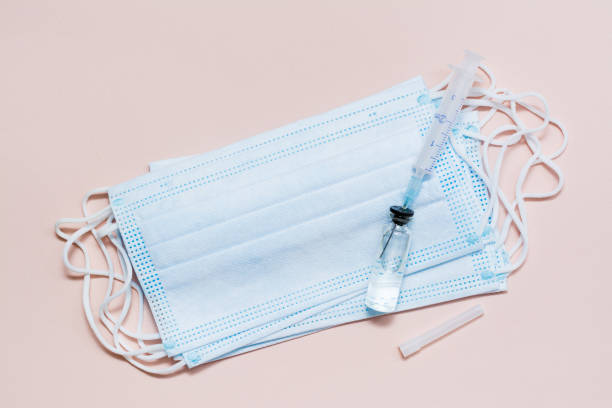 istock Vaccination and Immunization. Syringe needle inserted into a glass vial with vaccine on a face protective mask. Top view 1300865760