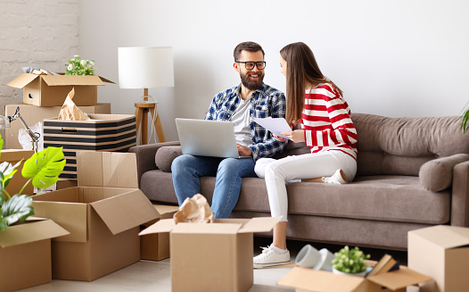 Happy young family couple with laptop and papers sitting on sofa in room with packed boxes and discussing insurance contract after relocation into new apartment