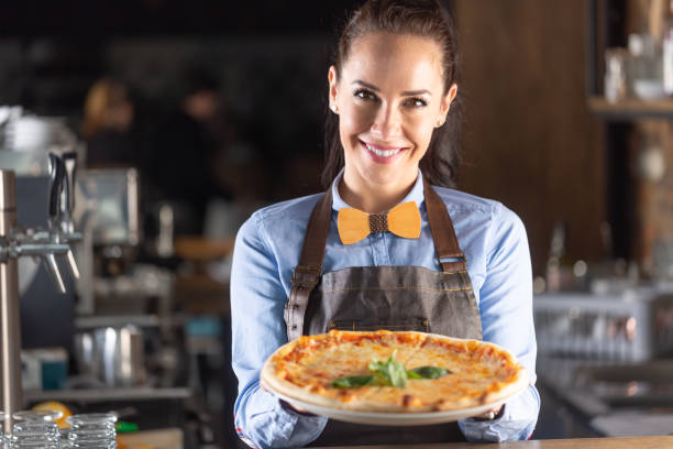 serveuse sourit tout en servant une grande pizza italienne authentique dans un restaurant. - pizza pizzeria restaurant waiter photos et images de collection