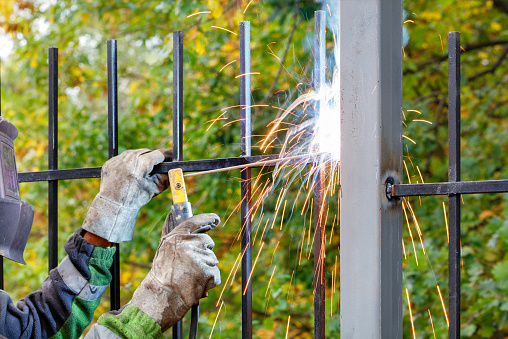 A welder in a protective helmet and gloves uses an electrode to weld an iron grate in a park area, bright sparks and blue smoke are flying. Selective focus, copy space.