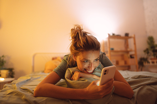 Photo of a young, smiling woman has a video call at night while lying on a bed in her bedroom