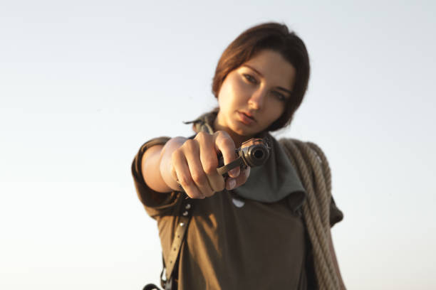 Woman standing holding a gun and rope outdoors in the desert. Portraint of a young brutal dangerous girl with weapon in confident pose. War action movie style beautiful armed female soldier dressed in stylish military clothes. Special forces serious woman aiming, ready for the attack. Focus on the handgun