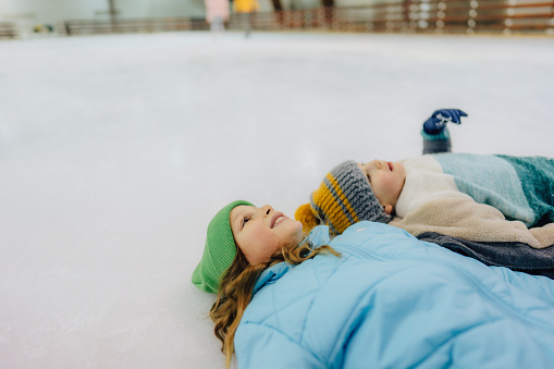 Photo of a little girl and her brother lying on the ice while having and ice-skating on the ice-rink.