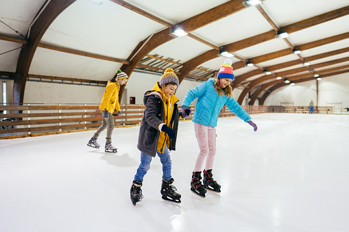 Photo of a little boy learning how to ice-skate on the ice rink, while his sister is holding his hand and guiding him.