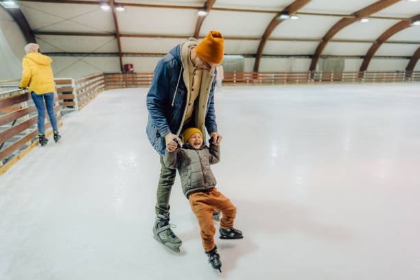Photo of a little boy learning how to ice-skate on the ice-skating rink, while his father is holding and showing him some basic tricks.