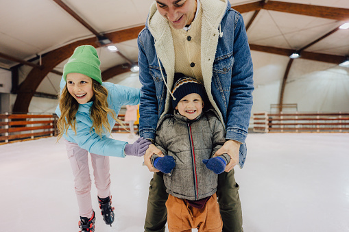 Photo of a cute little boy learning how to ice-skate on the ice rink, while his sister and father are holding and showing him some basic steps and tricks.