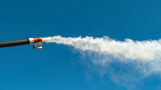 CLOSE UP, DOF: White smoke comes blasting out of a black fire extinguisher hose. Small portable fire extinguisher is set off outdoors on a sunny day. Detailed shot of a nozzle emitting white powder.