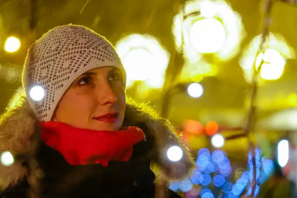 CLOSE UP, DOF: Beautiful Caucasian woman looks around the picturesque advent market. Gorgeous girl enjoying the sights of a Christmas market in big European city. Woman is enchanted by xmas lights.