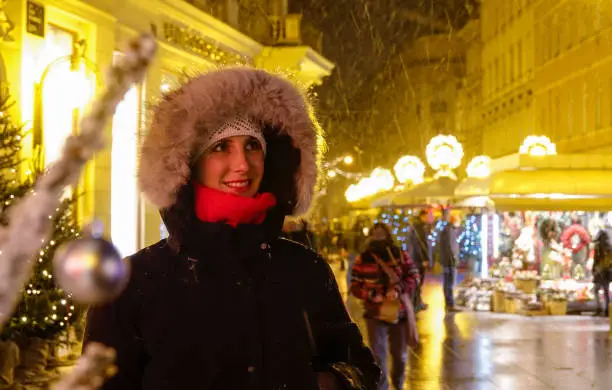 CLOSE UP, DOF: Smiling girl looks around the advent market on a snowy winter night in December. Beautiful Caucasian woman is enchanted by festive lights and market stalls of a Christmas market.