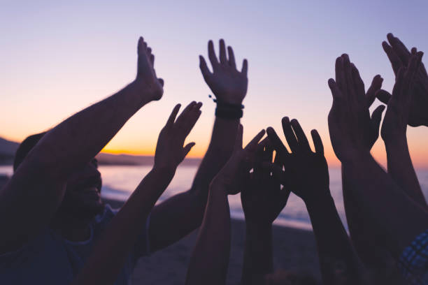 silhouette d’un groupe de personnes avec leurs mains levées au coucher du soleil ou au lever du soleil. - water human hand people women photos et images de collection