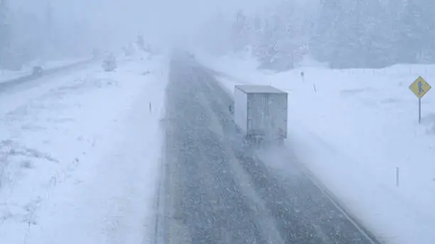 Cargo trucks and cars make their way through an intense blizzard along a dangerous country road leading through pine woods in Washington. Stunning shot of traffic moving through a bad snowstorm.