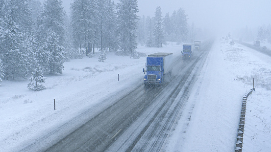 Freight trucks haul heavy cargo containers across the state of Washington and through a raging snowstorm. Cargo lorries navigate the slippery country road in low visibility during an intense blizzard.