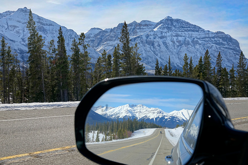 CLOSE UP, DOF: Side mirror of a moving car offers a picturesque view of the famous Icefields Parkway route and the stunning snowy mountains of Alberta. Car drives along scenic mountain road in Canada.