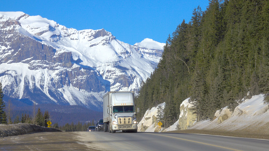 Big 18 wheeler and cars drive down the famous Icefields Parkway on a sunny spring day. Traffic moves along an interstate highway running under the snowy mountains. Road trip across Rocky Mountains.