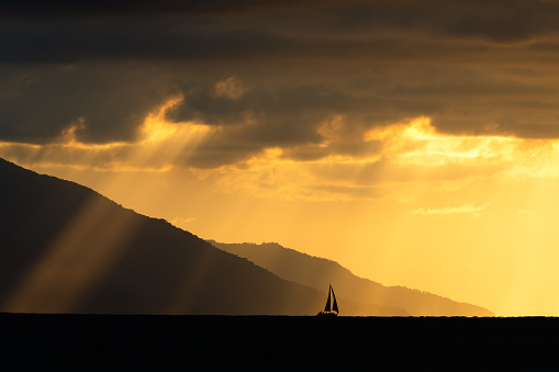 A Sailboat is Moving Along the Ocean as a Storm Looms and Sun Rays Break Through the Dark Clouds
