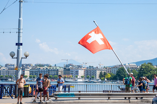 Picture of the Swiss flag in front of the Leman lake in Geneva, Switerland, in front of the Quai du Mont Blanc street and shritless male tourists. It is the main waterfront of the city of Geneve.