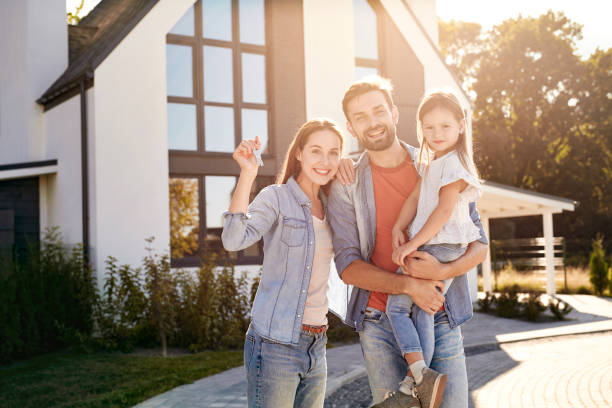 Urban Village. Family of three standing outdoors near house holding keys from new apartment in sunlight smiling cheerful stock photo