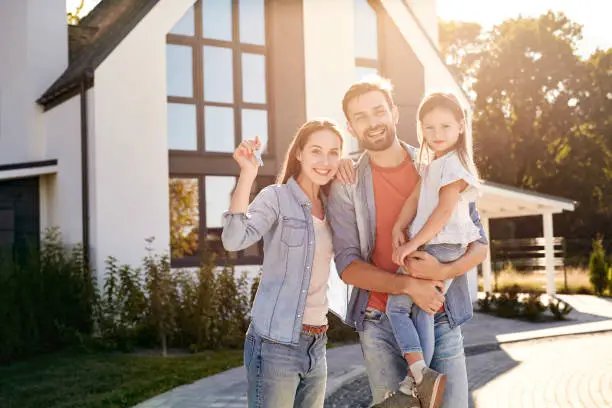 Family of three mother father and daughter standing together outdoors near house at urban village holding keys from new apartment in sunlight smiling cheerful