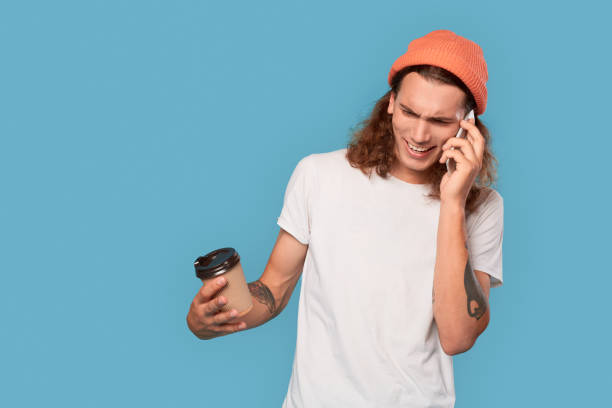 istock Freestyle. Curly-haired guy wearing hat standing isolated on blue with cup of coffee talking on smartphone wrinkling concerned 1300920017