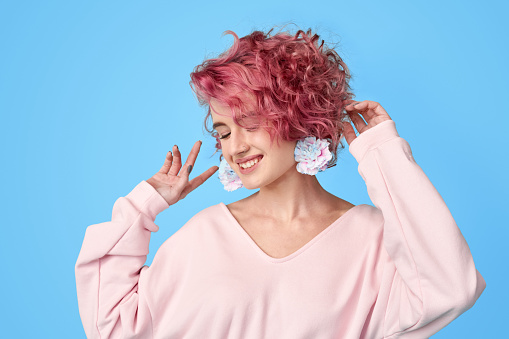 Portrait of dancing young girl with pink hair and carnation flower at earrings wearing in casual clothes, standing against blue background with copy space