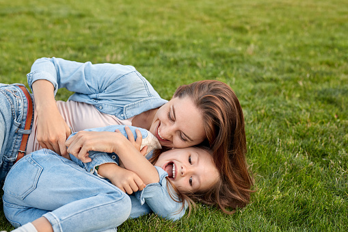 Young mother hugging daughter tickling her laughing playful lying together on lawn outdoors close-up