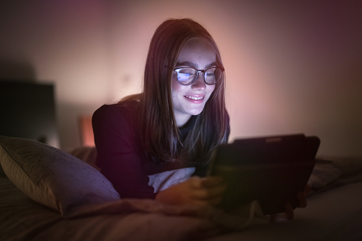 Smiling teenage woman lying in her bed in bedroom at night using her digital tablet computer. Having fun watching streaming videos - reading messages on her digital tablet. Natural Ambient Night Bedroom Light. Millenial Generation Modern Technology Daily Lifestyle.