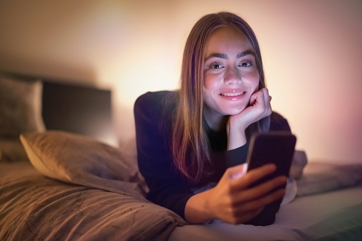 Smiling teenage woman relaxing in her bed in bedroom at night using her mobile phone. Lookinhg over to the camera, having fun watching streaming videos - reading messages on her smart phone. Natural Ambient Night Bedroom Light. Millenial Generation Modern Technology Daily Lifestyle.