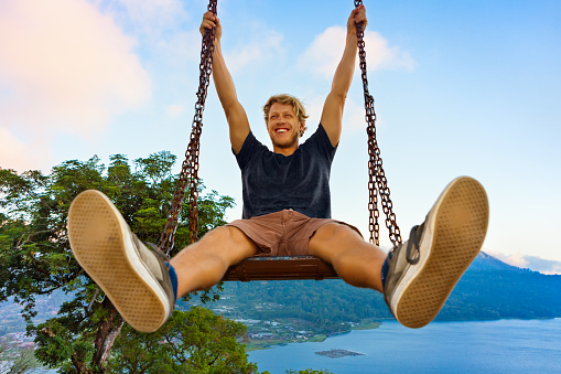 Summer vacation. Young man sit on tree rope swing with amazing jungle view. Swinging high with fun above tropical lake. Buyan lake is popular travel destinations in Bali island, Indonesia