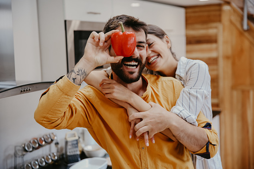 Portrait of a playful couple making goofy faces and playing with pepper while preparing food together in a modern kitchen.